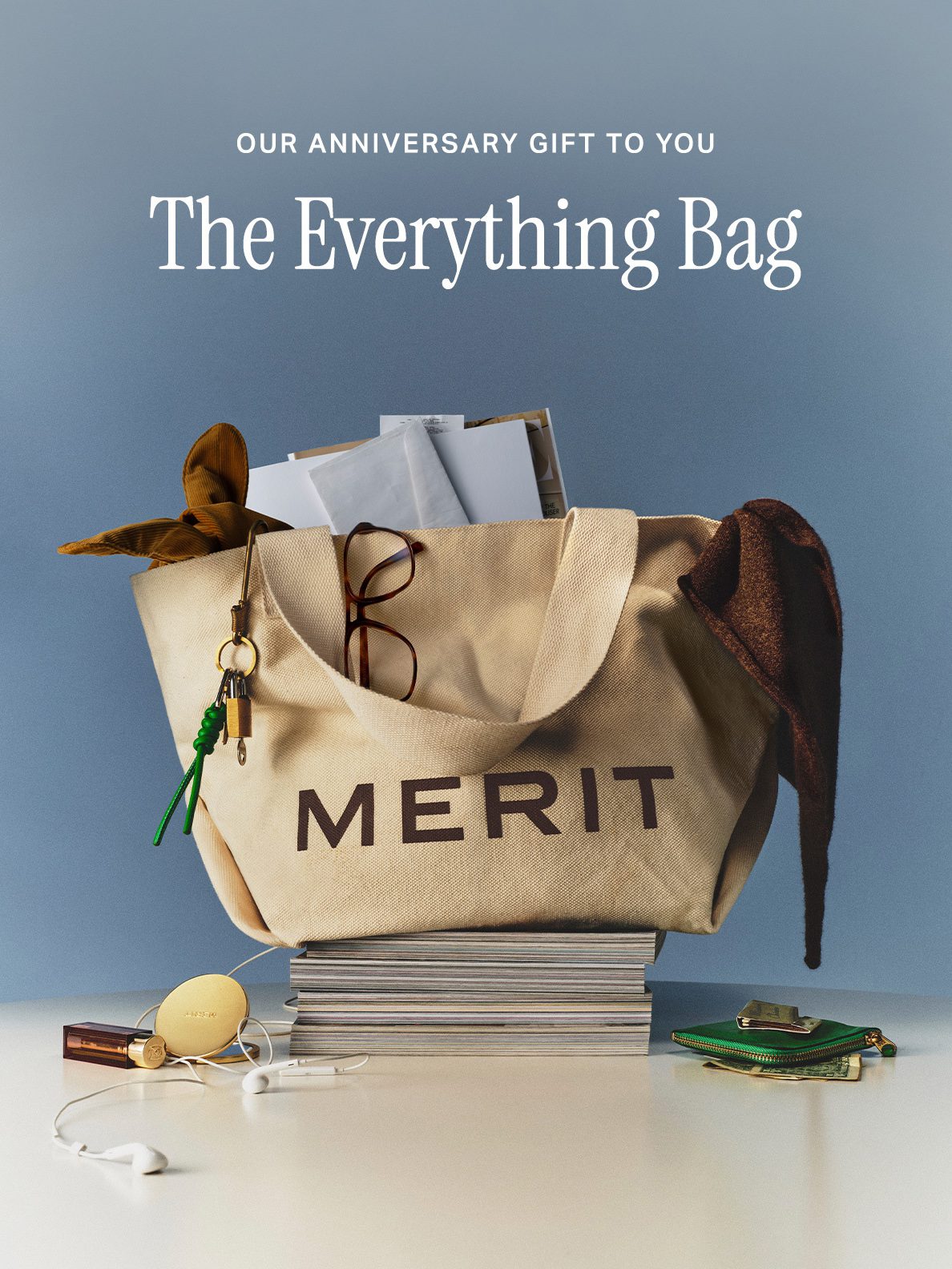 A cream MERIT tote bag on a pedestal against a pale blue background, with “The Everything Bag” text above it.